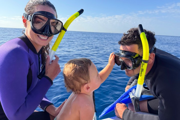 Two snorkelers and a child near the ocean, smiling on a sunny day.