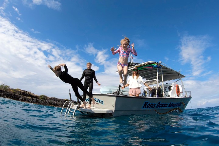 People jump off a boat into the ocean under a cloudy sky.