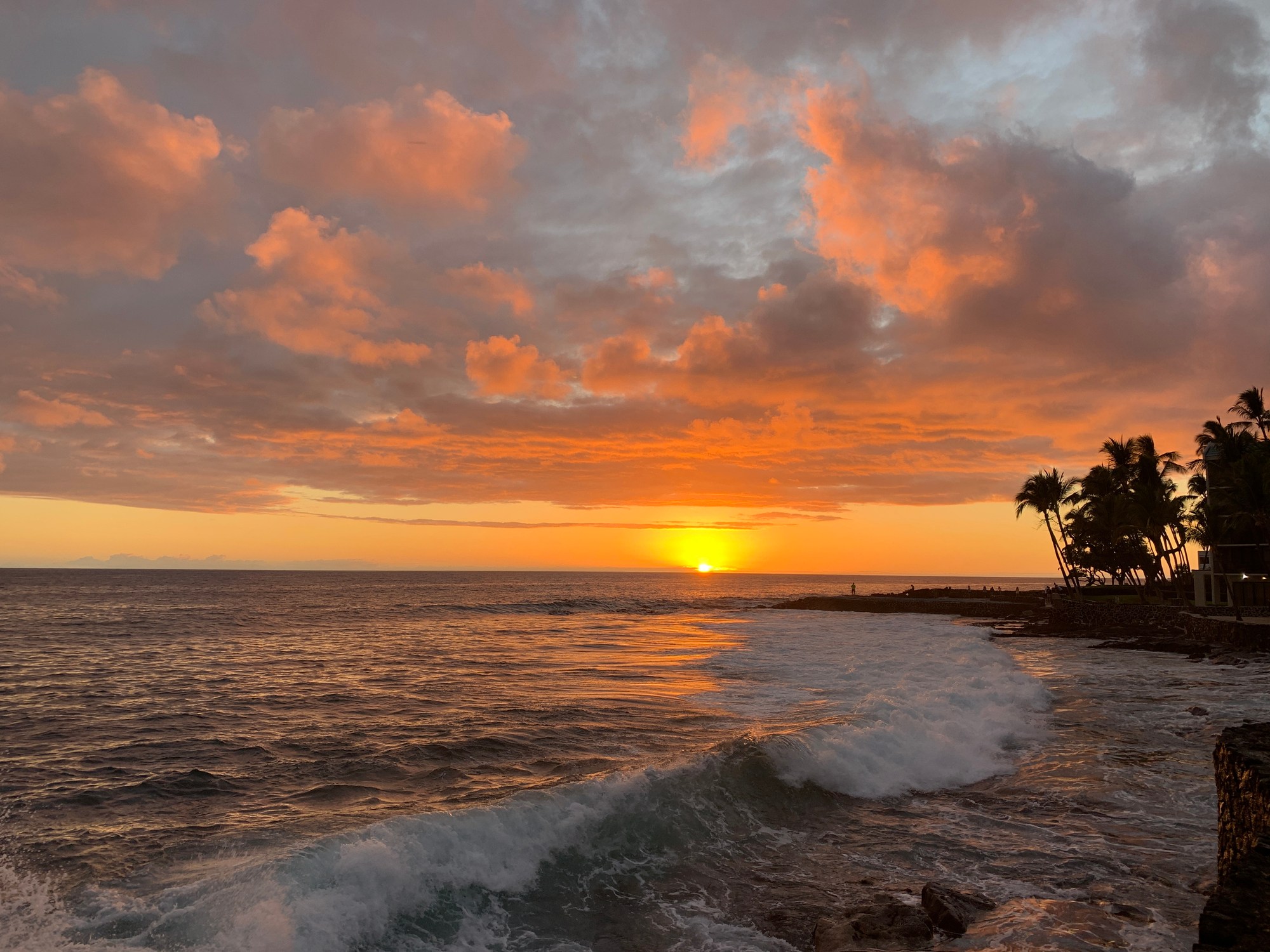 Stunning sunset in Kona, Hawaii, viewed from the shore. The sky turns orange above a calm ocean.